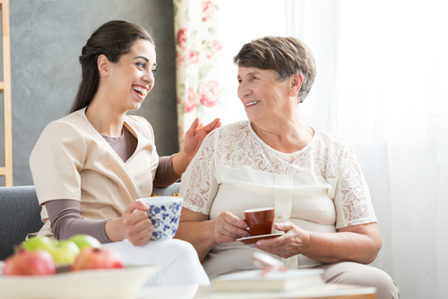 Nurse and senior woman having fun together while sitting on sofa and drinking tea